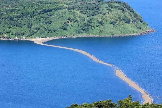 A thin sand spit curving from a green headland out into the sea